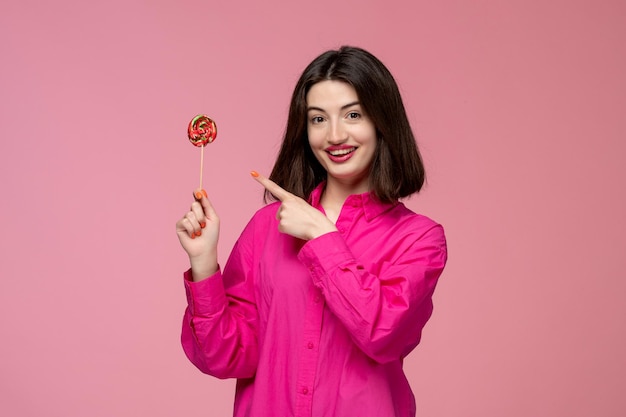 Nettes Mädchen hübsches entzückendes Mädchen im rosa Hemd mit dem roten Lippenstift, der auf Lutscher zeigt