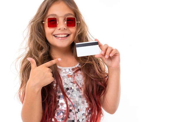 Nettes Mädchen in der quadratischen roten Sonnenbrille, die Plastikbankkarte hält und auf sie lokalisiert zeigt