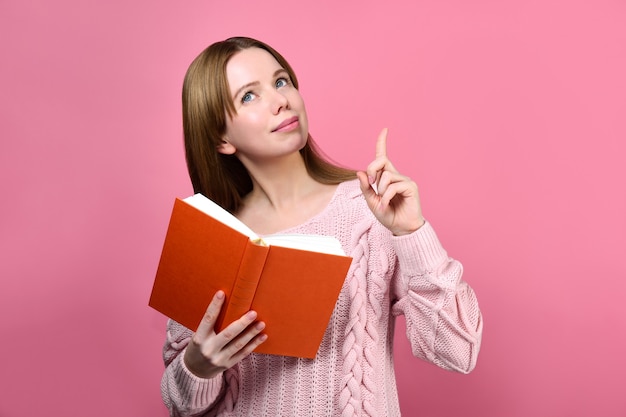 Nettes Mädchen in einem rosa Pullover hält ein Buch und hob den Zeigefinger.