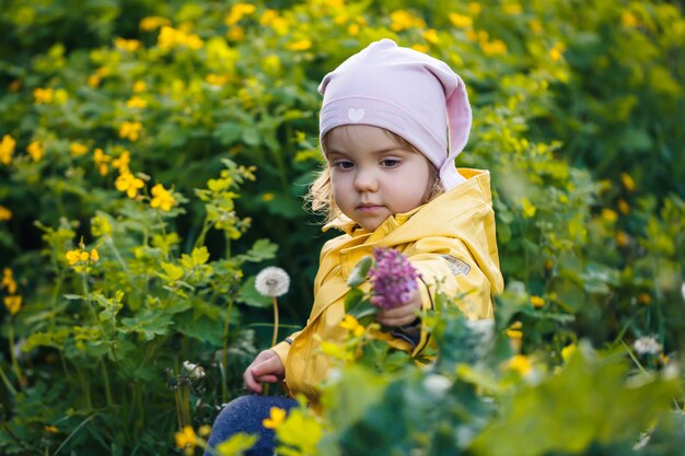 Nettes Mädchen in einer gelben Jacke sammelt Blumen auf einer Wiese. Ein Kind stand im frischen grünen Gras zwischen den Frühlingsblumen. Ein kleines Kind macht einen Strauß aus den ersten wilden gelben Blumen