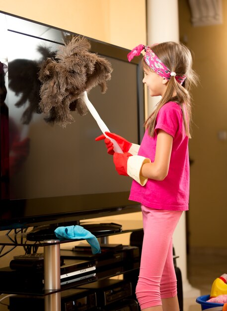 Nettes mädchen in gummihandschuhen, das großen fernseher mit bürste vom staub säubert