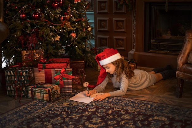 Nettes Mädchen in Weihnachtsmütze, das einen Brief an den Weihnachtsmann im weihnachtlich dekorierten Wohnzimmer schreibt