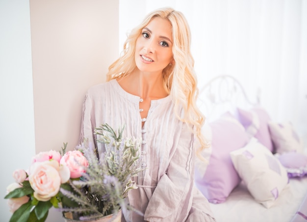 Nettes Mädchen mit blonden Haaren im lila Kleid und in den weißen Socken sitzt auf Bett im hellen Schlafzimmer