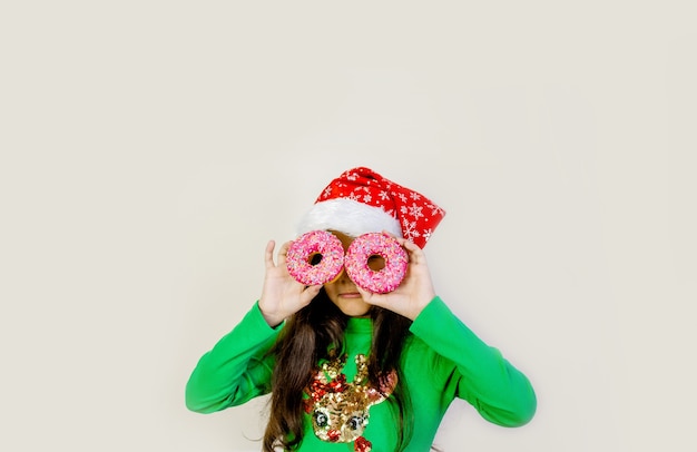 Nettes Mädchen mit dunklem Haar in der Weihnachtsmütze, die lächelt, Spaß hat und durch zwei Donuts auf ihren Augen schaut.