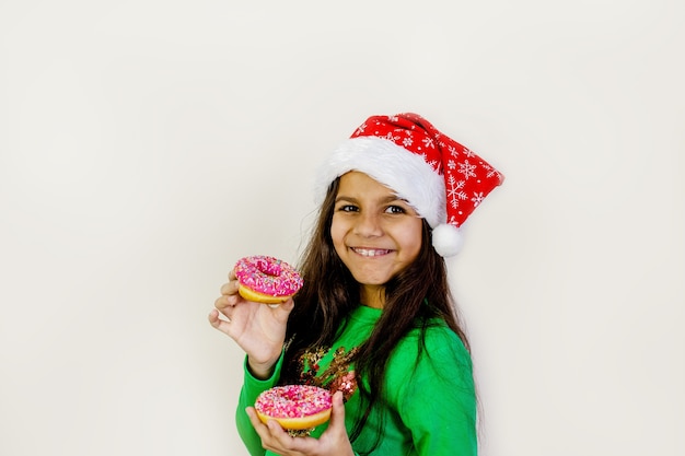 Nettes Mädchen mit dunklem Haar in der Weihnachtsmütze, die lächelt, Spaß hat und zwei Donuts auf ihren Augen schmeckt.