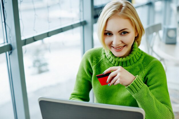 Nettes Mädchen mit hellem Haar, das grünen Pullover trägt, sitzt im Café mit Laptop und Kreditkarte, Porträt, freiberufliches Konzept, Online-Einkauf.