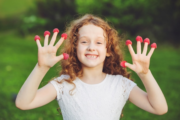 Nettes Mädchen mit Himbeeren auf ihren Fingern, die ihre Zähne zeigen.