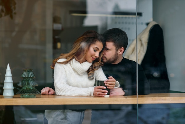 Nettes Mädchen mit ihrem Freund, der innerhalb eines Cafés sitzt und heißen wohlriechenden Kaffee trinkt, während draußen kaltes Wetter ist. Liebe und Romantik.