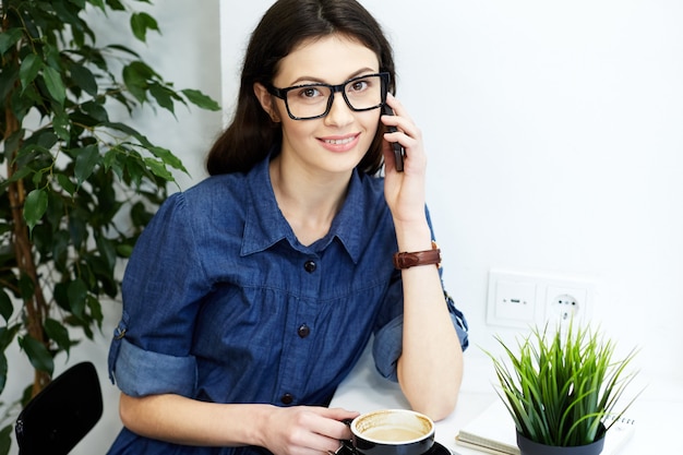 Nettes Mädchen mit schwarzen Haaren, die blaues gestreiftes Hemd und Brillen tragen, die im Café mit Tasse Kaffee, freiberufliches Konzept, Porträt, lächelnd, am Handy sprechen, Kamera betrachten tragen.