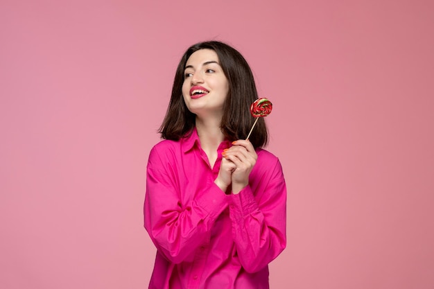 Nettes Mädchen reizendes junges Brunettemädchen im rosa Hemd mit dem roten Lippenstift, der roten Lutscher hält