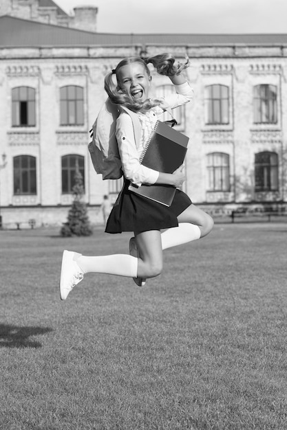 Nettes Mädchen springt mit Notizbuch glückliches Vorschulmädchen mit Buch im Schulhof zurück zur Schule fleißiges Kind trägt Rucksackkonzept der Bildung Frühlingsferienzeit Sommerferien