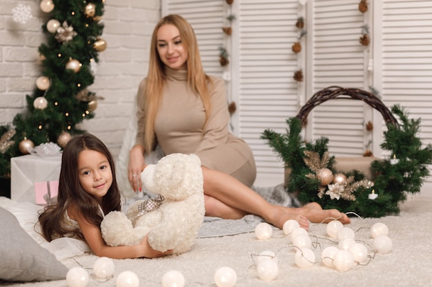 Nettes Mädchen und ihre Mutter spielen mit Teddybär in der Nähe des Weihnachtsbaumes zu Hause