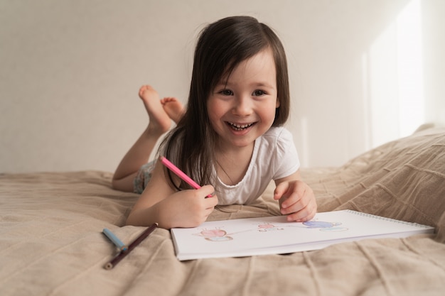 Nettes Mädchen zeichnet eine Bleistiftzeichnung. Ein Kind lernt, Menschen zu zeichnen. Das Kind liegt auf dem Bett und zeichnet mit einem Bleistift