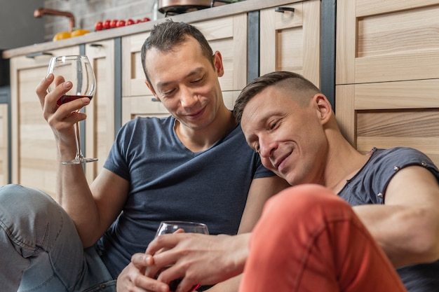 Nettes männliches schwules Paar verbringt Zeit zusammen. Sie trinken Wein auf dem Küchenboden sitzend