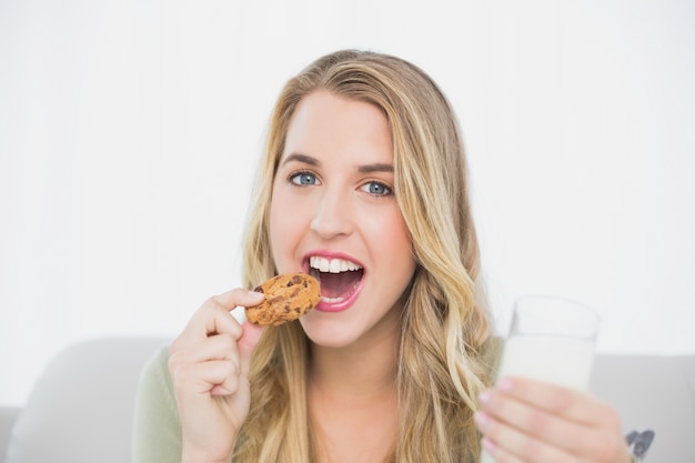 Nettes nettes blondes essendes Plätzchen mit der Milch, die auf gemütlichem Sofa sitzt