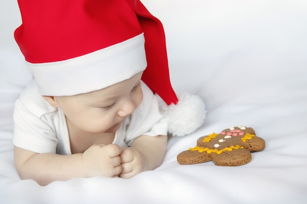 Nettes Neugeborenes im Weihnachtsmann-Weihnachtsmannkostüm mit Lüge auf weißem Blatt und Lebkuchen betrachten