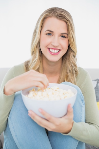 Nettes recht blondes essendes Popcorn, das auf gemütlichem Sofa sitzt