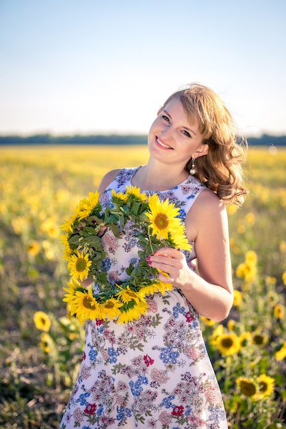 Nettes rothaariges lächelndes Mädchen auf dem Gebiet voller Sonnenblumen, Kranz aus Sonnenblumen.