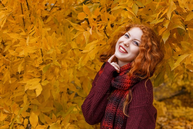 Nettes rothaariges Mädchen mit einem Herbstbaum mit einem Blatt in ihrer Hand lächelt und verzieht das Gesicht