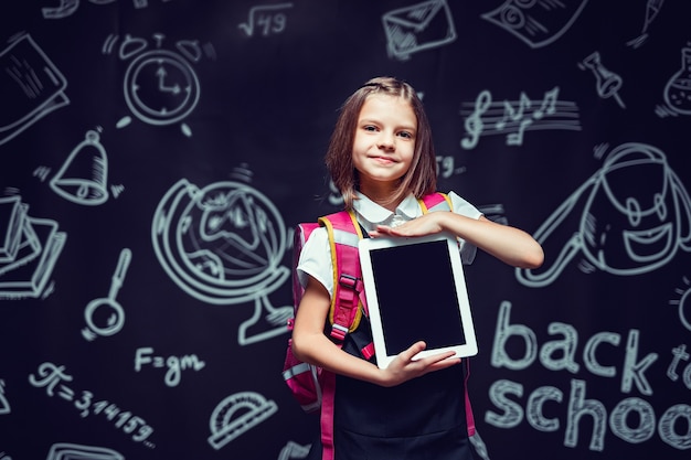 Nettes Schulmädchen, das sich darauf vorbereitet, mit Rucksack zur Schule zu gehen, der das Tablet zurück zum Schulkonzept zeigt