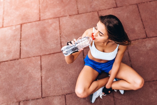 Nettes sportliches Mädchen, das nach Training im Freien mit Flasche Wasser in den Händen sich entspannt