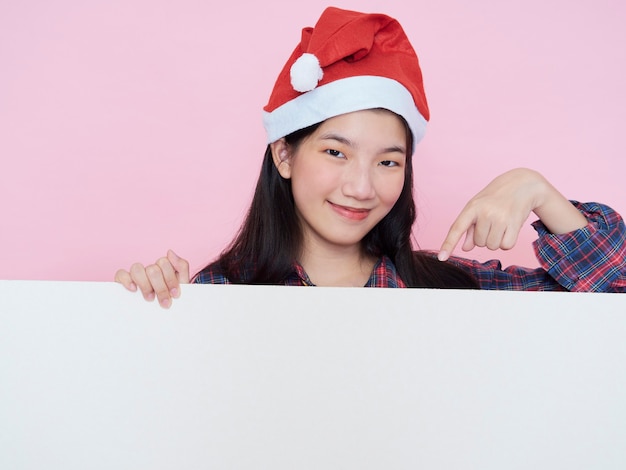 Nettes Teenager-Mädchen in der Weihnachtsmütze, die Finger auf leerem Plakat zeigt, während auf rosa Hintergrund steht.