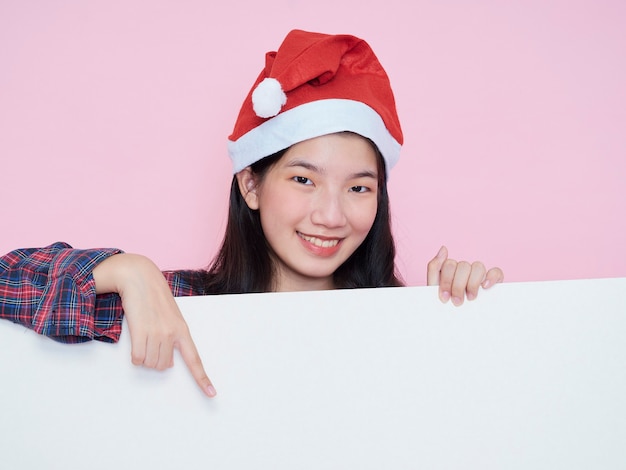 Nettes Teenager-Mädchen in der Weihnachtsmütze, die Finger auf leerem Plakat zeigt, während auf rosa Hintergrund steht.