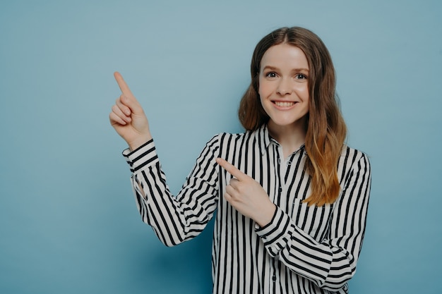 Nettes Teenager-Mädchen in gestreiftem Hemd mit glänzendem Lächeln, das mit den Zeigefingern auf Kopienraum für Werbung zeigt, positives weibliches Werbeprodukt im Stehen einzeln auf blauem Hintergrund