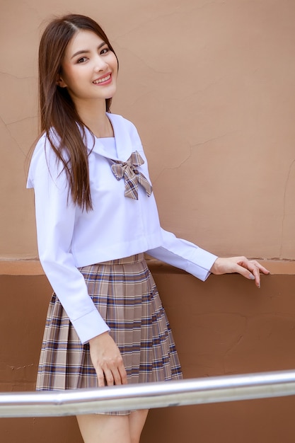 Nettes und junges Mädchen, das japanische, koreanische Schulmädchenuniform trägt und mit Spaß und glücklich vor dem Schulgebäude vor der Kamera posiert.