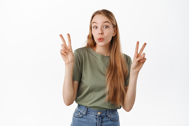 Nettes und lustiges blondes Mädchen zeigt Friedens-V-Zeichen und verzogene Lippen albern und steht glücklich in Sommer-T-Shirt und Jeans gegen weiße Wand