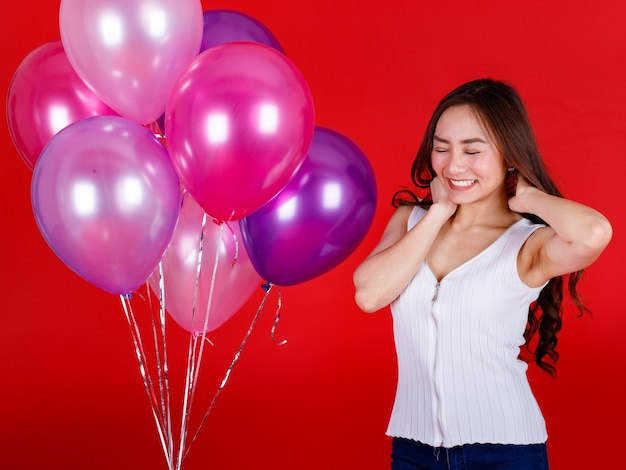 Nettes und schönes asiatisches Mädchen, das mit bunten Luftballons mit einem lustigen und glücklichen Lächeln auf rotem Hintergrund stinkt und spielt, Studiolichtschießen. Festival und feiern Konzept.