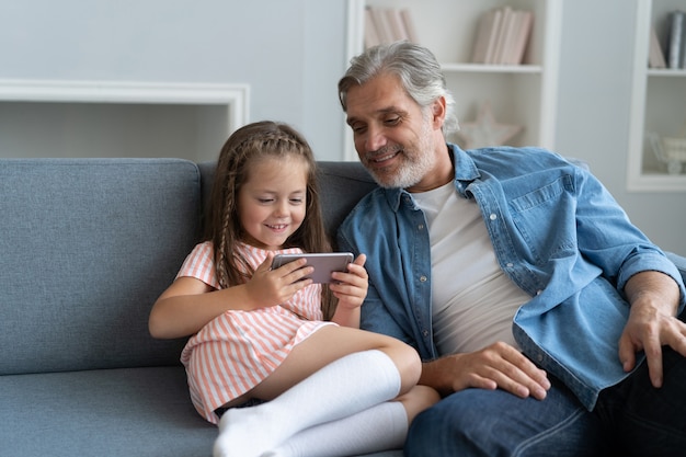 Nettes Vorschulmädchen hilft Vater, etwas auf dem Smartphone zu zeigen, intelligente kleine Tochter und Papa sitzen auf der Couch und halten Handy