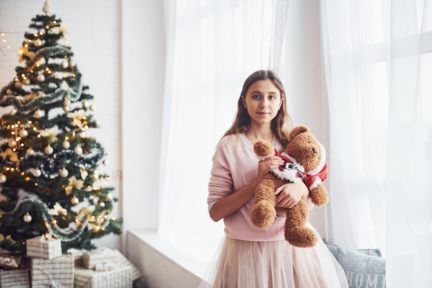 Nettes weibliches jugendlich, das zuhause im wohnzimmer zur ferienzeit mit teddybär steht.