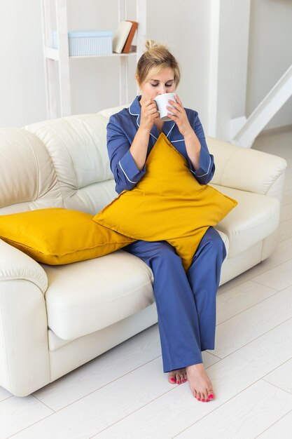 Nettes weibliches Model im Pyjama genießt morgens eine angenehme Frau, die auf dem Sofa sitzt und Tee oder Kaffee trinkt