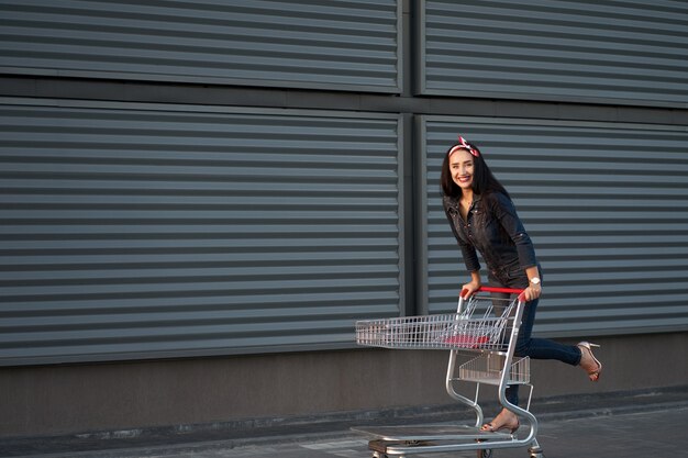 Neue moderne Kollektion. Porträt lustiges lächelndes modisches brünettes Mädchen mit langen Haaren, die trendige Jeansoveralls tragen, die einen Einkaufswagen reiten. Schuss auf grauem Stadtwandhintergrund.