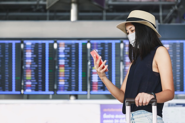 Neue normale Reisende asiatische Frau mit Maske und Gepäck unter Verwendung des Mobiltelefons im Terminalflughafen Thailand