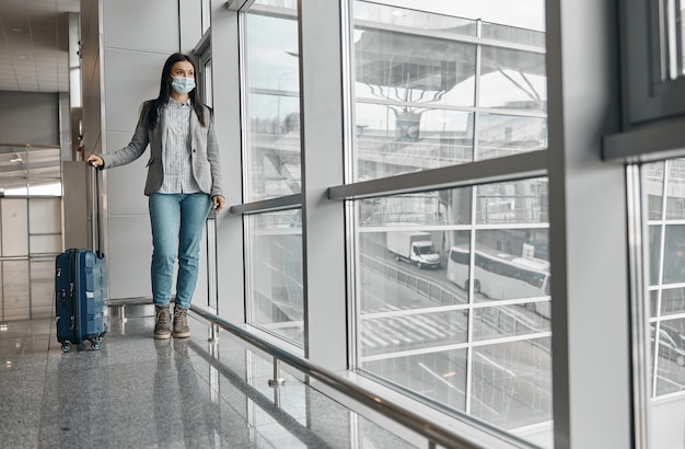 Neue Ordnung an Flughäfen. Frau mit Maske im Terminal mit Gepäck allein in der Nähe von Panoramafenstern