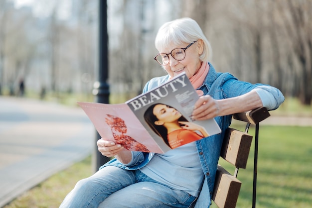 Neue Trends. Lächelnde blonde Frau, die eine Zeitschrift liest, während sie auf der Bank sitzt
