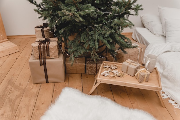 Neues 2023 2024-jähriges Weihnachtszimmer in beigen Farbtönen, dekoriert für Feiertage. Viele Geschenke unter dem Weihnachtsbaum und Glühbirnen. Beiges Tablett mit Neujahrssüßigkeiten