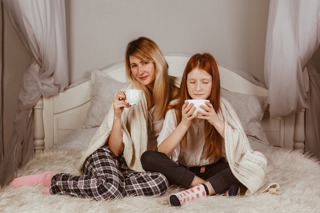 Neues Jahr 2020. Mutter und Ingwerhaartochter sitzen auf dem Bett und trinken Kakao mit Schaum. Mit einer Decke bedeckt, umarmen.