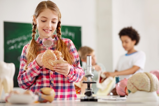 Neues Wissen. Fröhliches positives Mädchen, das Lupe hält, während eine Lektion über Biologie hat