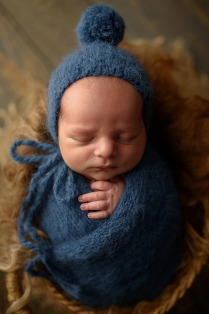 Neugeborener Junge in einer blauen Strickmütze und Wicklung schläft in einem Korb auf einem hölzernen Hintergrund.