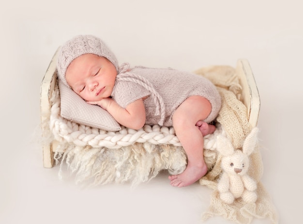 Neugeborenes Baby trägt gestricktes beige Kostüm und Hut, das auf seinem Bauch in einem kleinen Bett schläft und winzige Hände unter seinen Wangen hält. Entzückendes Nickerchen für Kleinkinder