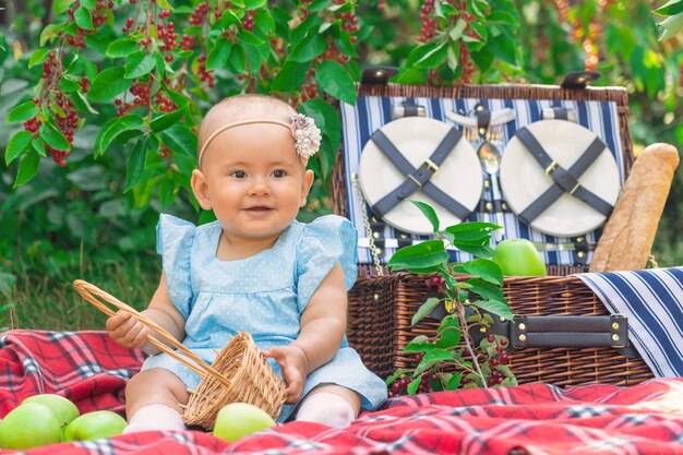Neugeborenes Mädchen in einem blauen Kleid auf einem Picknick. Ein 9-12 Monate Kleinkind mit einem leeren Korb und grünen Äpfeln herum