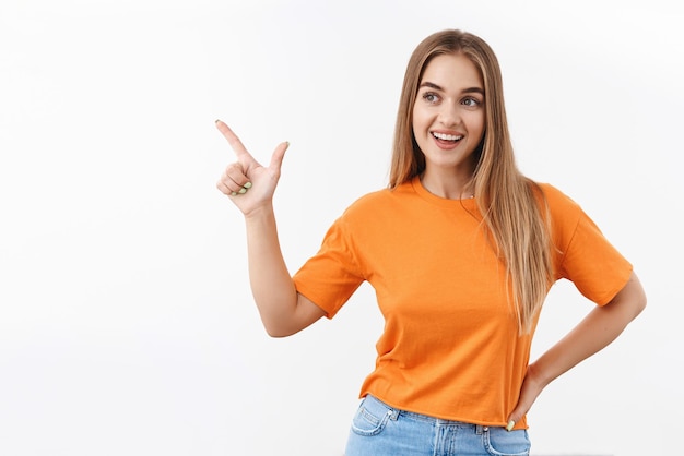 Neugierig wunderschönes, stilvolles blondes Mädchen, Teenager in orangefarbenem T-Shirt, Auswahl treffen, Artikel im Laden auswählen, auf die obere linke Ecke zeigen und schauen