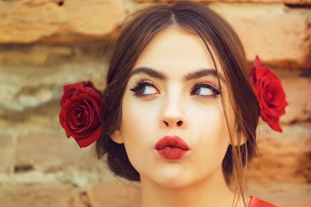 Neugierige Frau oder süßes Mädchen, junges Modemodell mit roten Lippen, Make-up und frischen Rosen, Blumen im brünetten Haar, modische Frisur, die draußen auf alter Backsteinmauer aufwirft. Schönheits- und Friseursalon