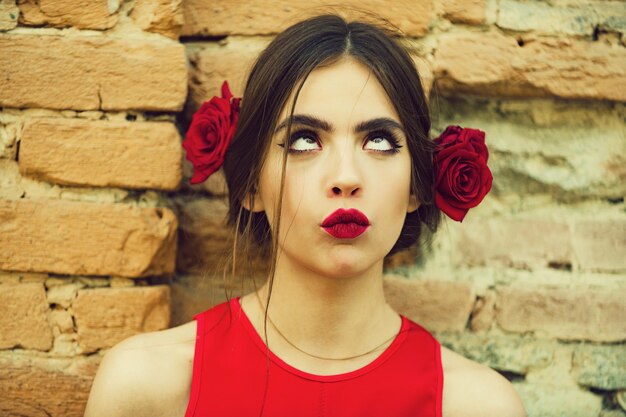 Neugierige Frau oder süßes Mädchen, junges Modemodell mit roten Lippen, Make-up und frischen Rosen, Blumen im brünetten Haar, modische Frisur, die draußen auf alter Backsteinmauer posiert. Schönheits- und Friseursalon
