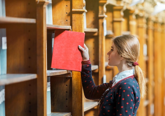 Neugierige Frau wählt Buch in der Nähe von Bibliotheksbücherregalen mit rotem Buch aus