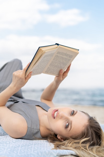 Neugierige junge blonde Frau, die ein Buch hält, während sie am Strand liegt