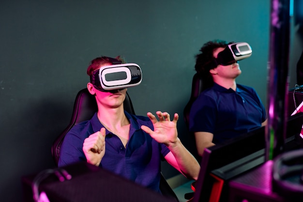 Neugierige junge Männer in denselben T-Shirts sitzen im dunklen E-Sport-Club und verwenden Virtual-Reality-Simulatoren, während sie Netzwerk-Videospiele spielen